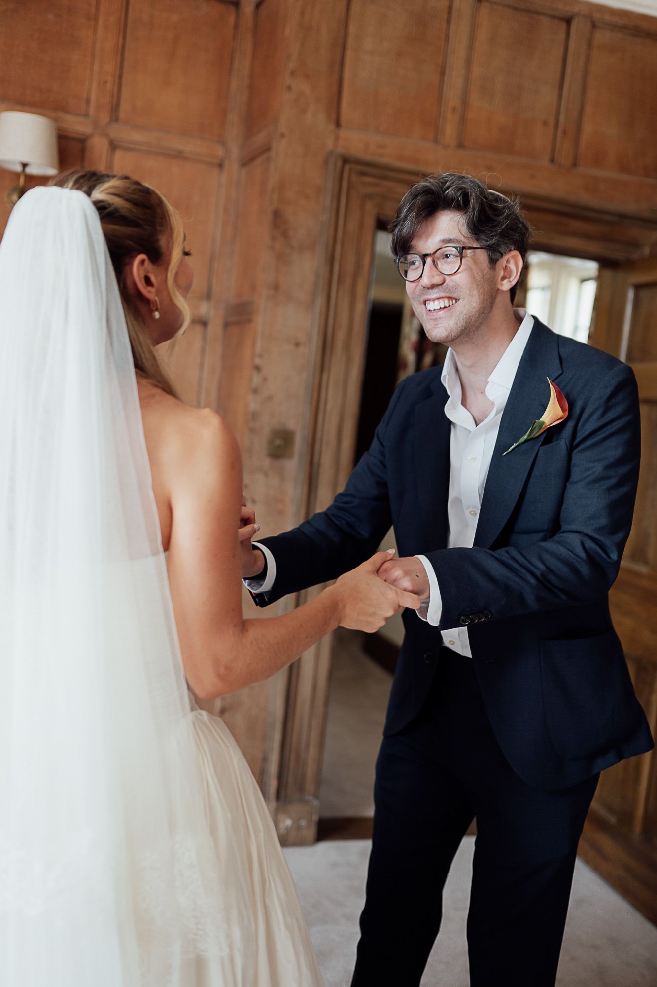 Smiling groom in navy suit holds bride's hands during first look moment in elegant wood-paneled room.