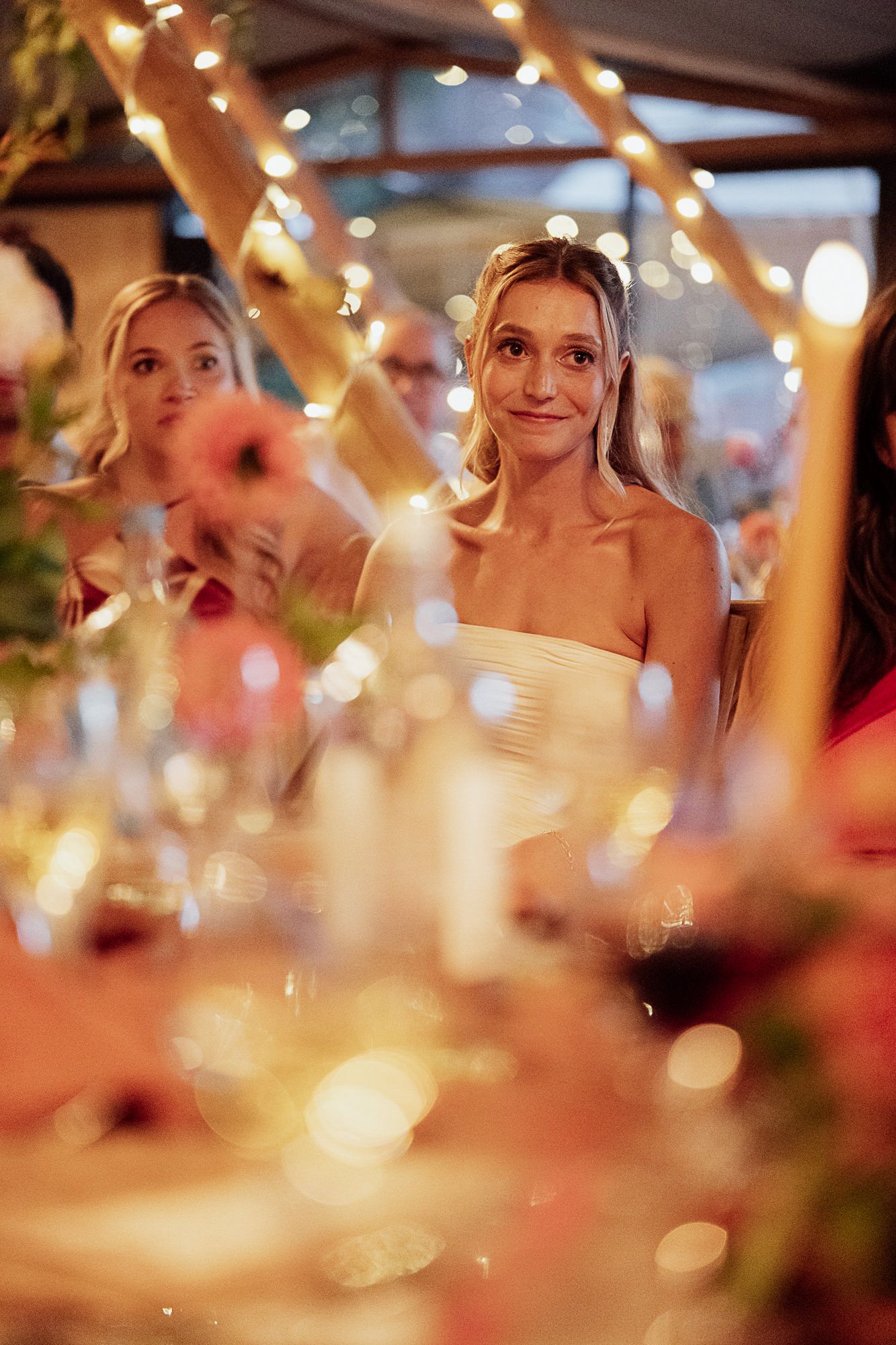 A beautiful bride in a strapless white gown listens at a candlelit wedding reception with fairy lights.