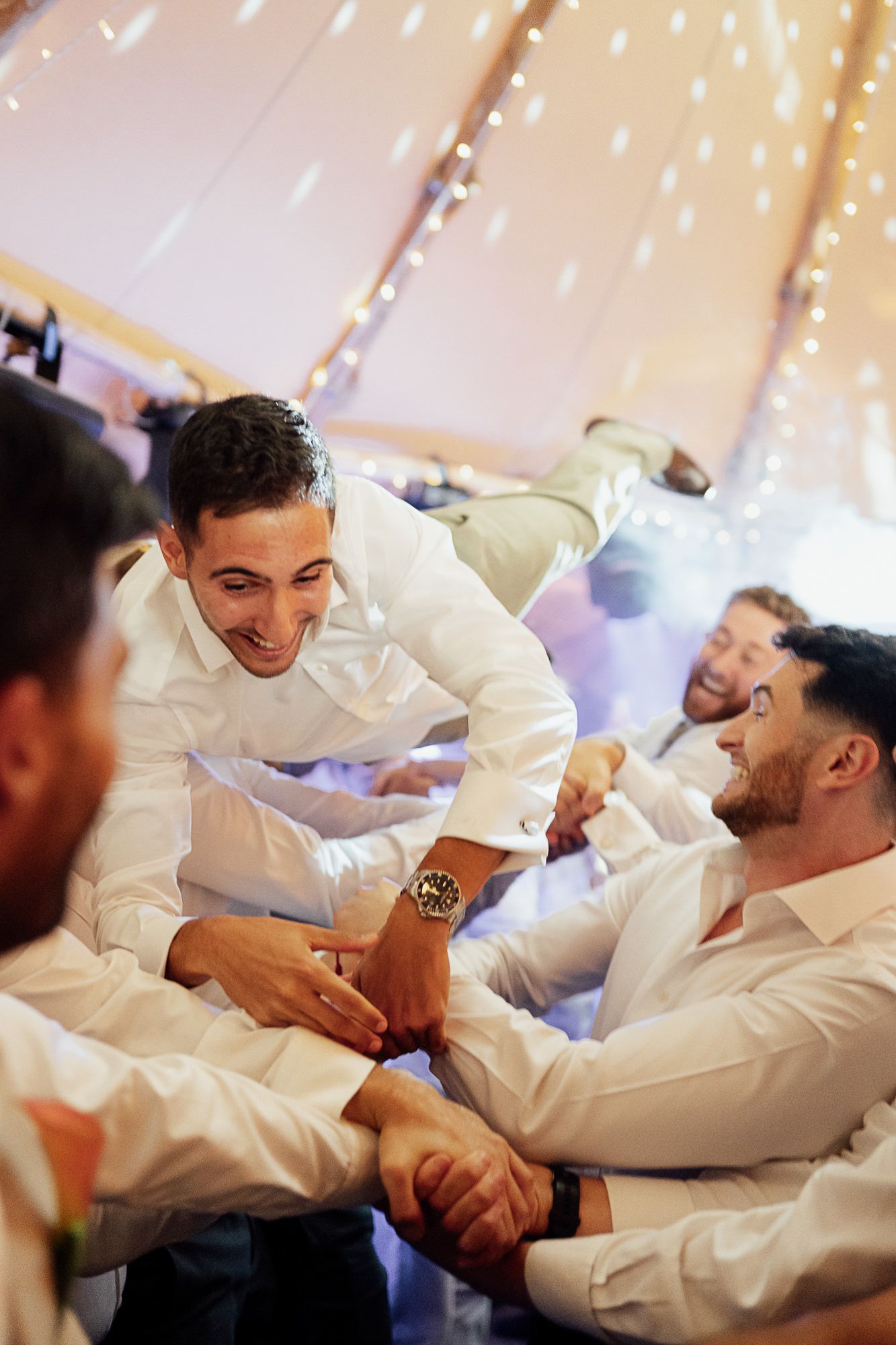 Groomsmen in white shirts laughing and celebrating together under fairy lights at a wedding reception tent.