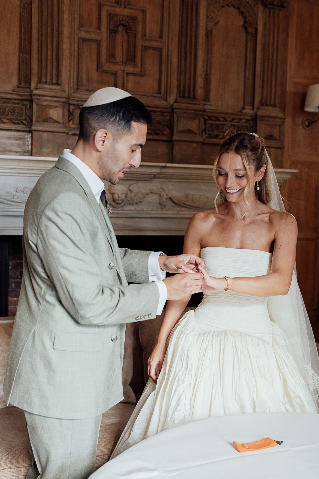 Groom placing wedding ring on bride's finger during Jewish ceremony in elegant wood-paneled room.