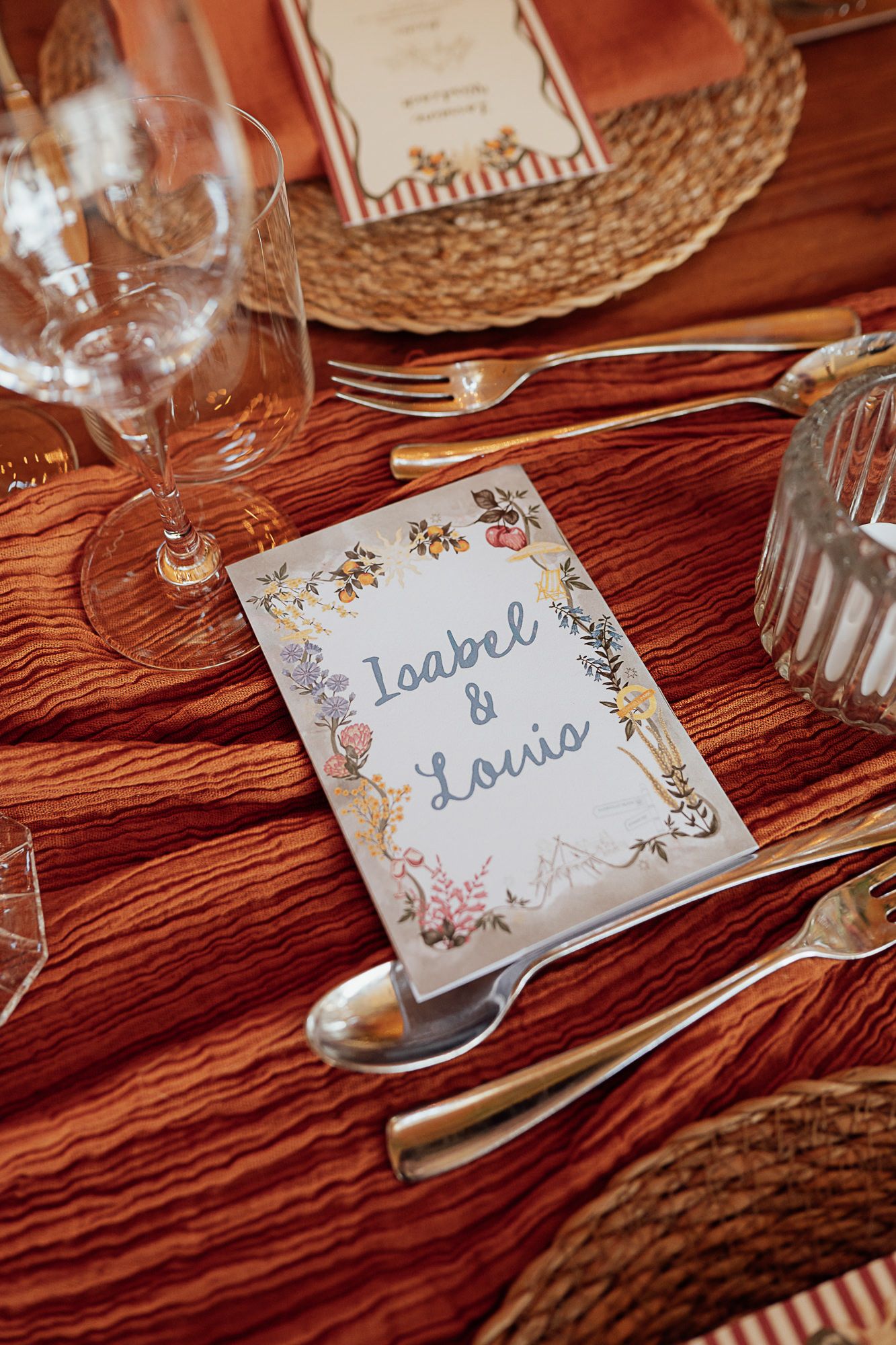 Elegant wedding place setting with Isabel & Louis card, gold cutlery, woven charger, and rust-colored linen tablecloth.