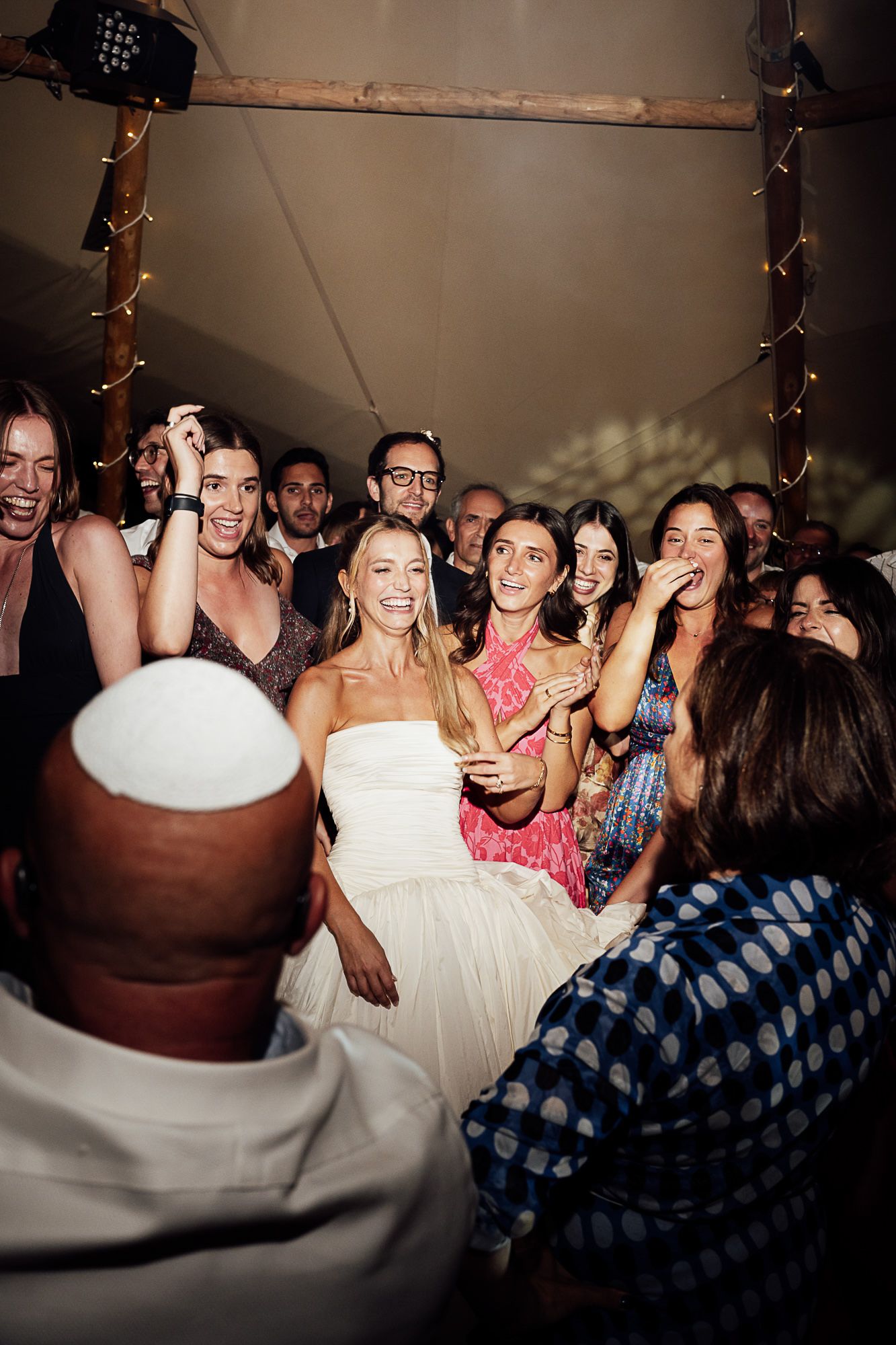 Bride in white dress laughing joyfully with wedding guests under a tent decorated with fairy lights.