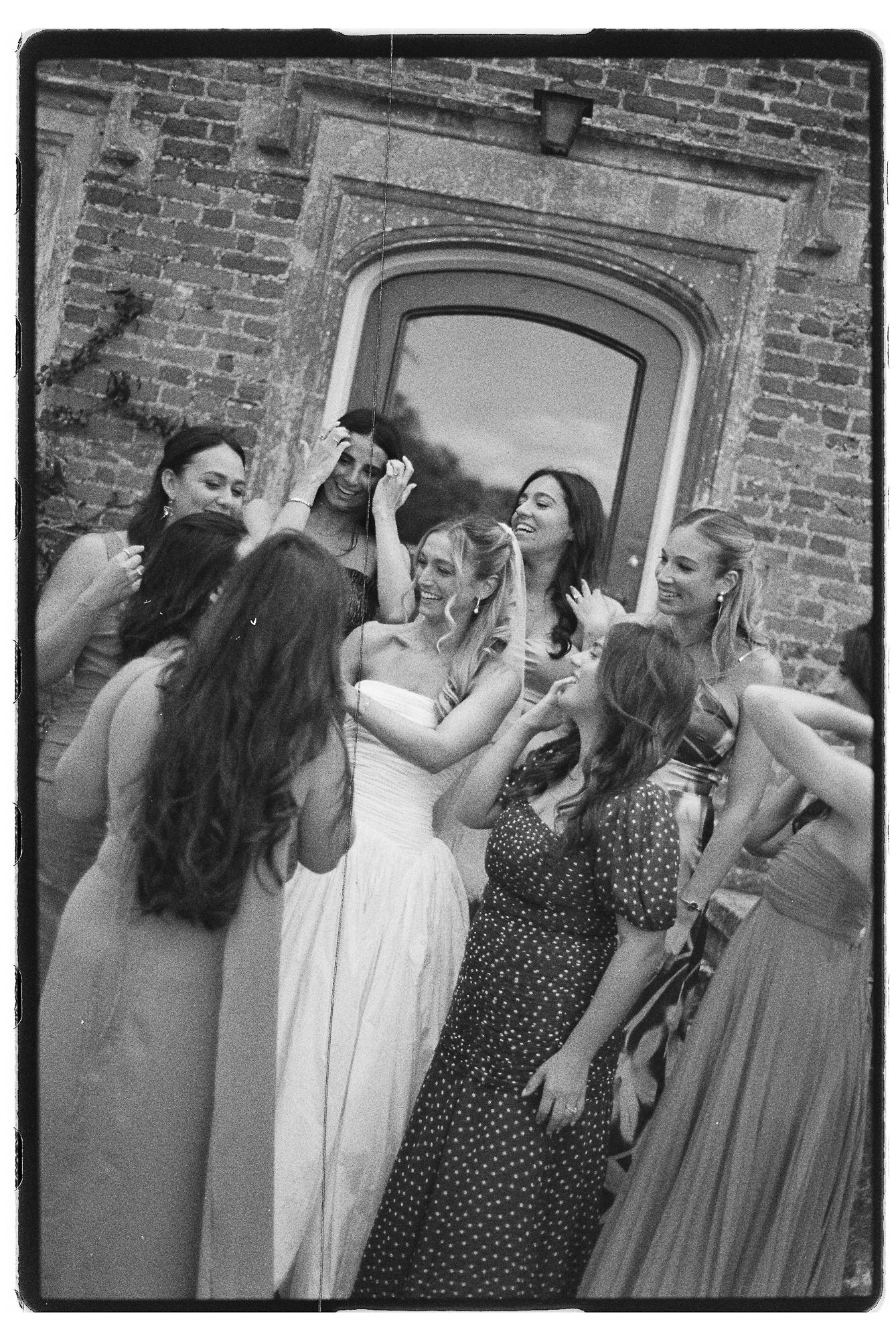 Black and white photo of a bride in white gown surrounded by laughing bridesmaids outside a brick building.