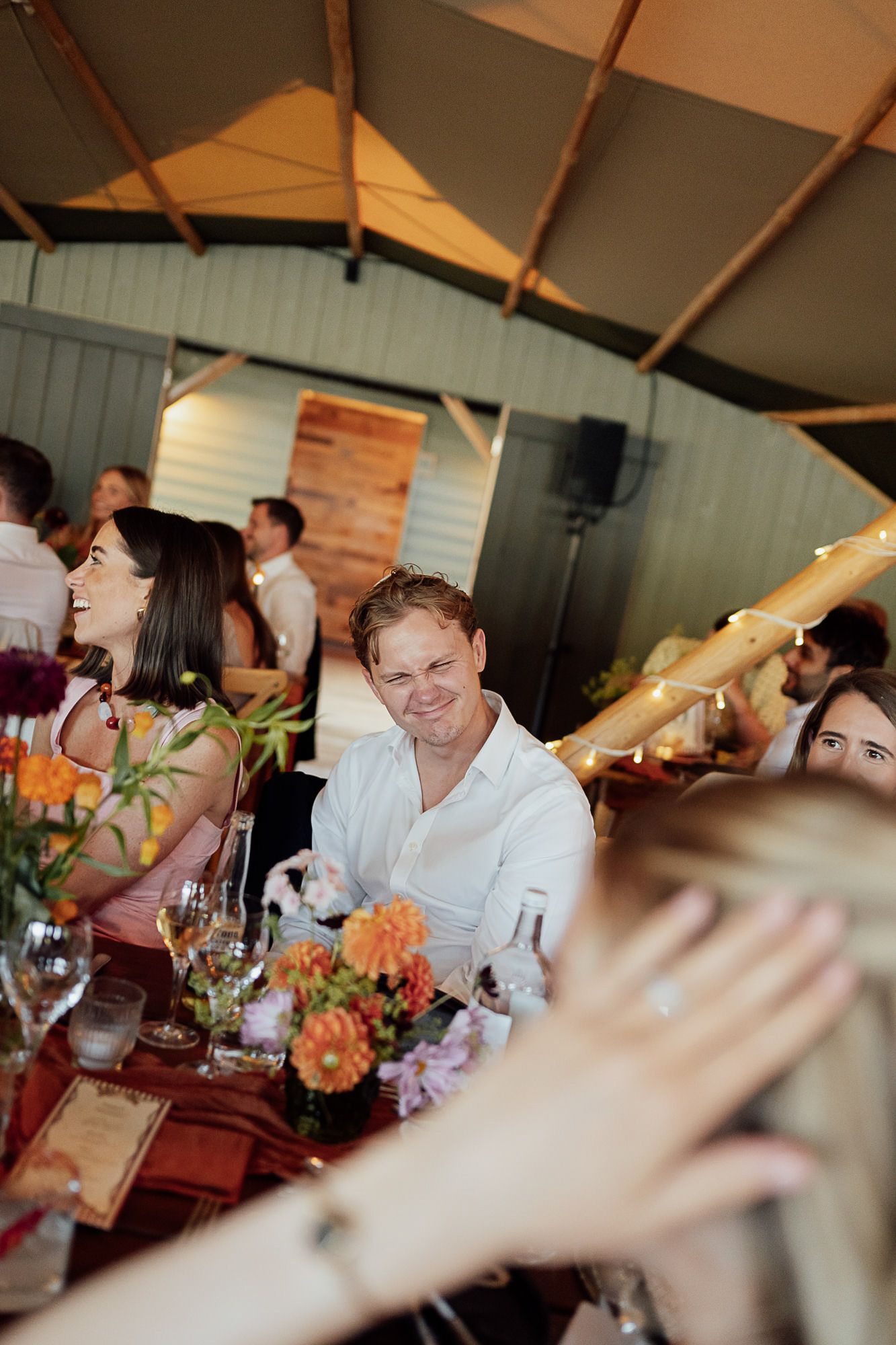 Guests seated at a decorated wedding reception table with colorful floral centerpieces inside a tent venue.