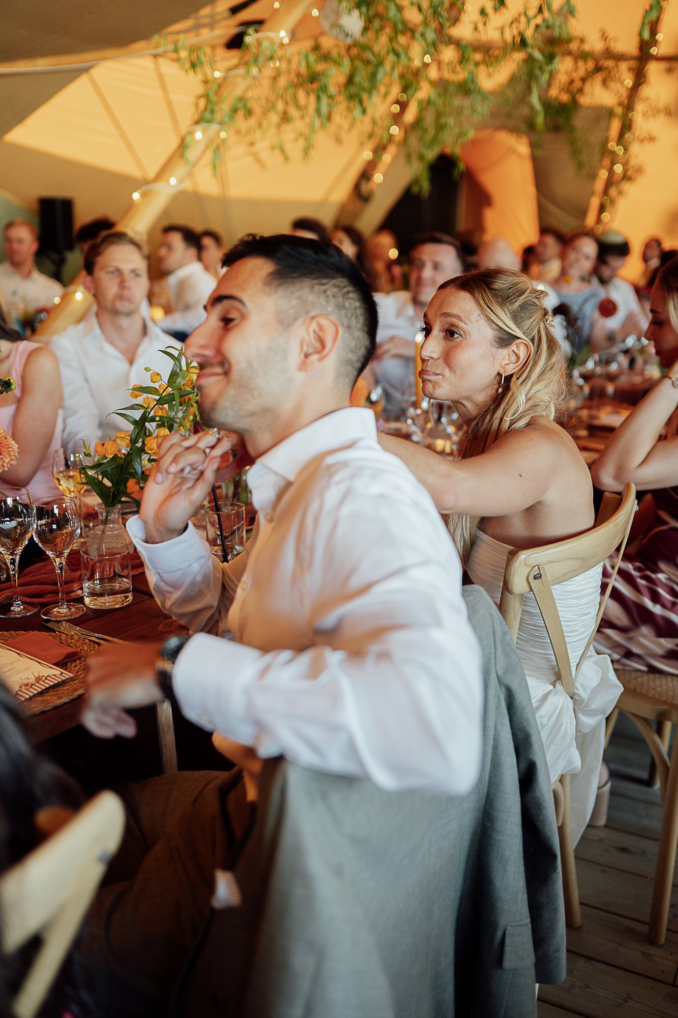 Guests seated at decorated tables during an elegant outdoor wedding reception under a tent with string lights.