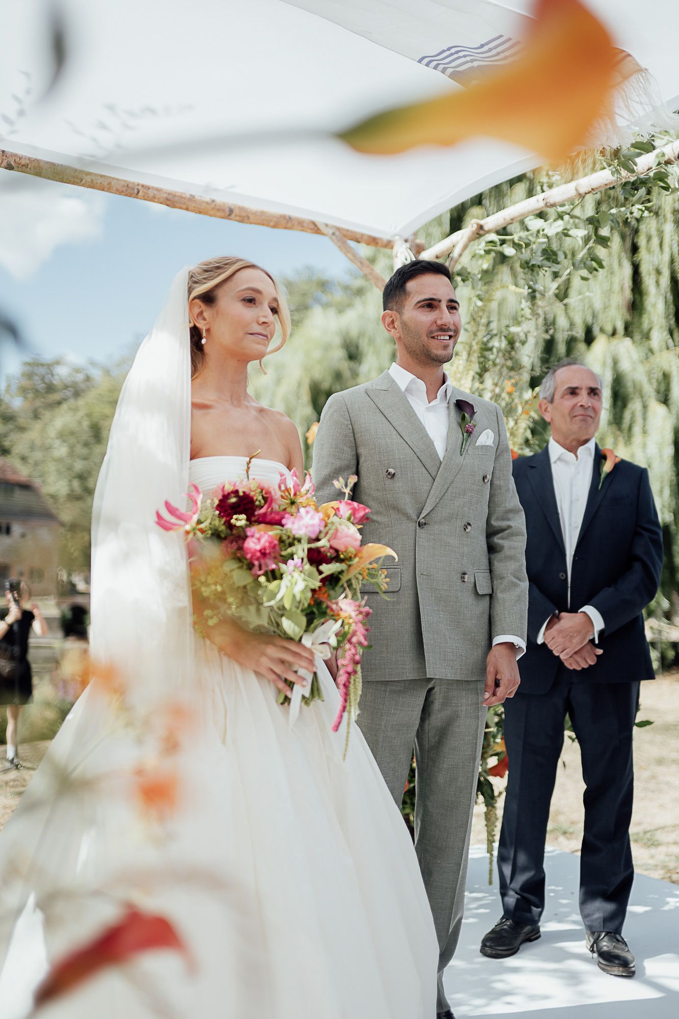 Bride and groom stand at outdoor altar under floral arch, bride holds colorful bouquet, officiant behind them.