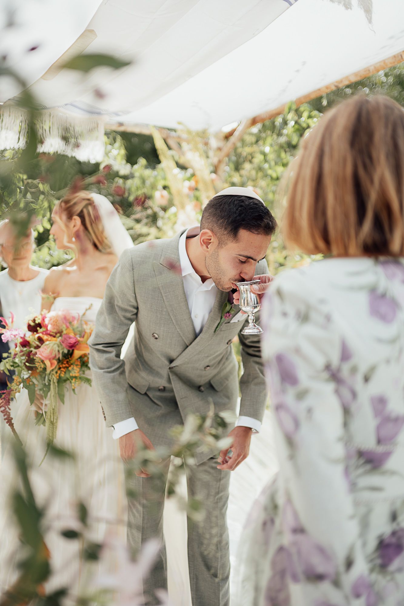 Groom in gray suit drinking from ceremonial cup under floral chuppah at outdoor Jewish wedding ceremony.