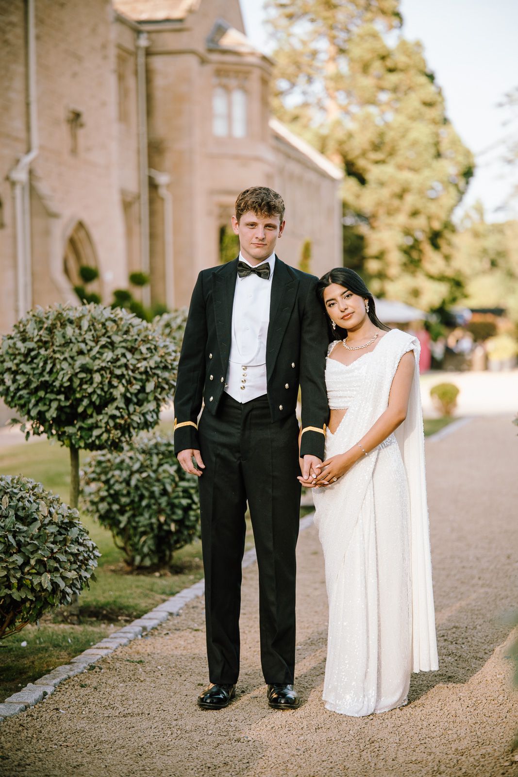 Elegant couple in formal attire, groom in black tuxedo and bride in white saree, posing outdoors near a grand building.