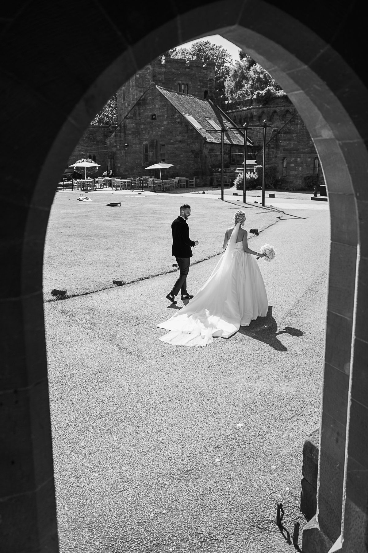 Black and white photo of bride and groom walking through a stone arch at a historic venue on their wedding day.