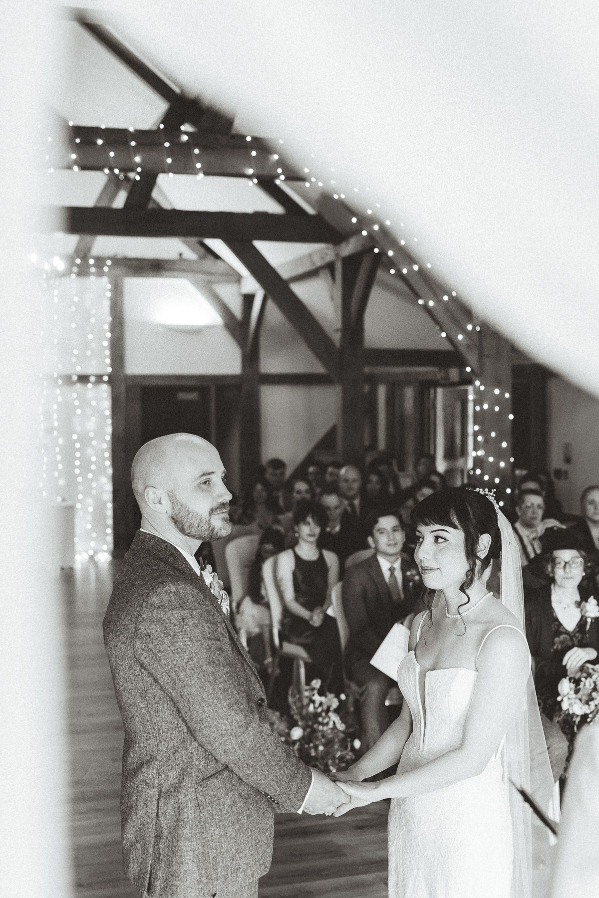 Black and white wedding ceremony photo showing a couple exchanging vows under rustic wooden beams with string lights.