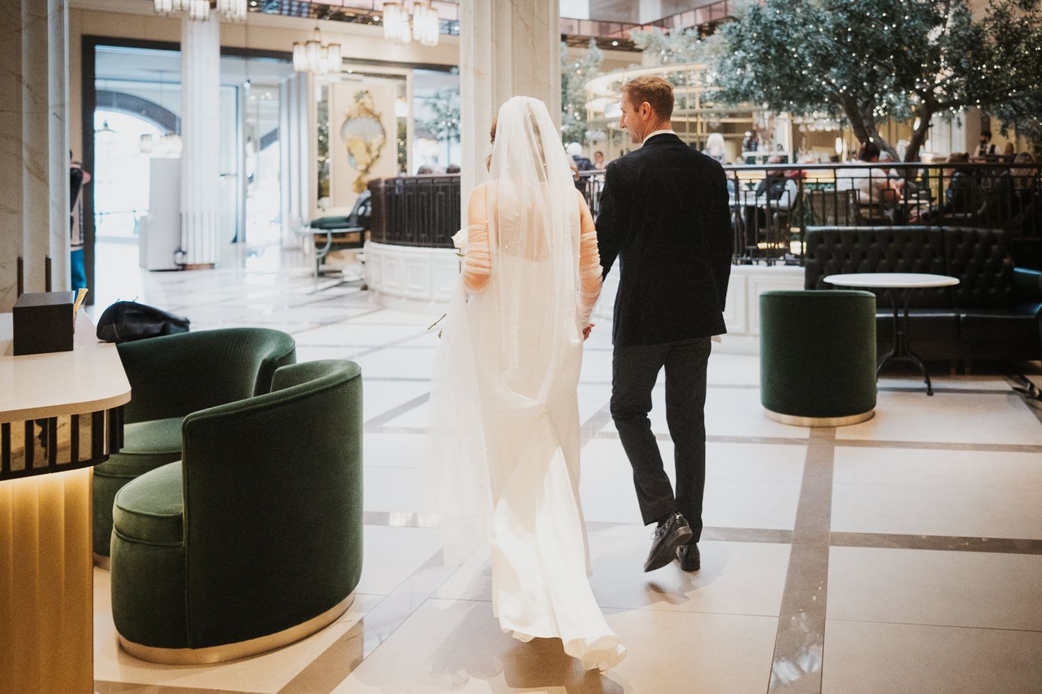 A couple in wedding attire walks through an elegant venue with dark green velvet chairs and modern decor.