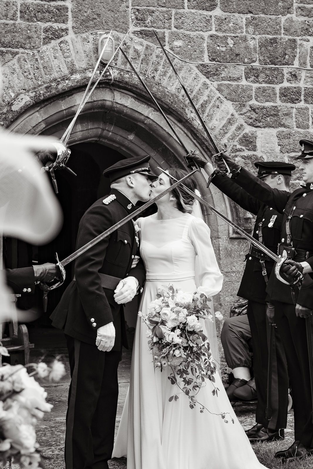 Military officers with swords form an arch for a newlywed couple outside a stone church after their wedding ceremony.