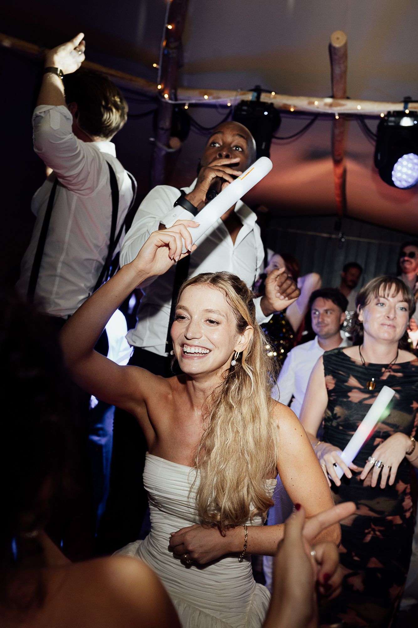 Bride dancing joyfully at wedding reception holding glow stick, surrounded by guests celebrating on dance floor.