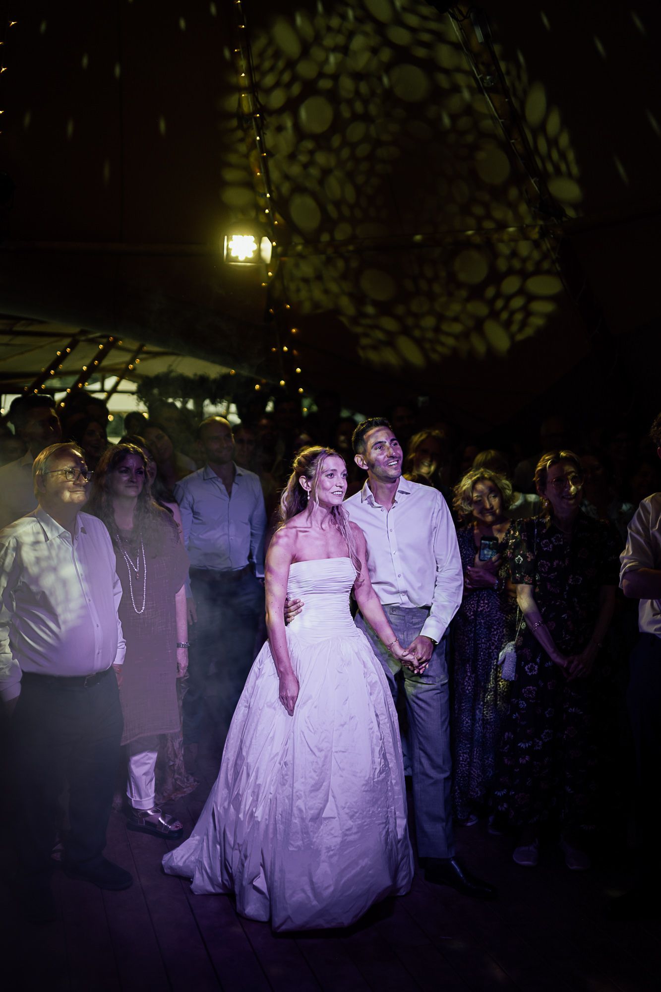 Bride in white gown and groom hold hands during first dance at elegant wedding reception with decorative lighting.