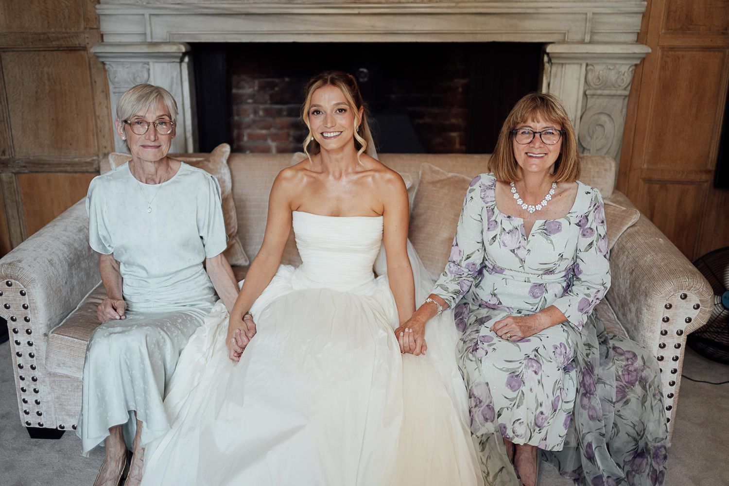 Bride in white gown sits between two older women on a cream sofa in front of a stone fireplace.