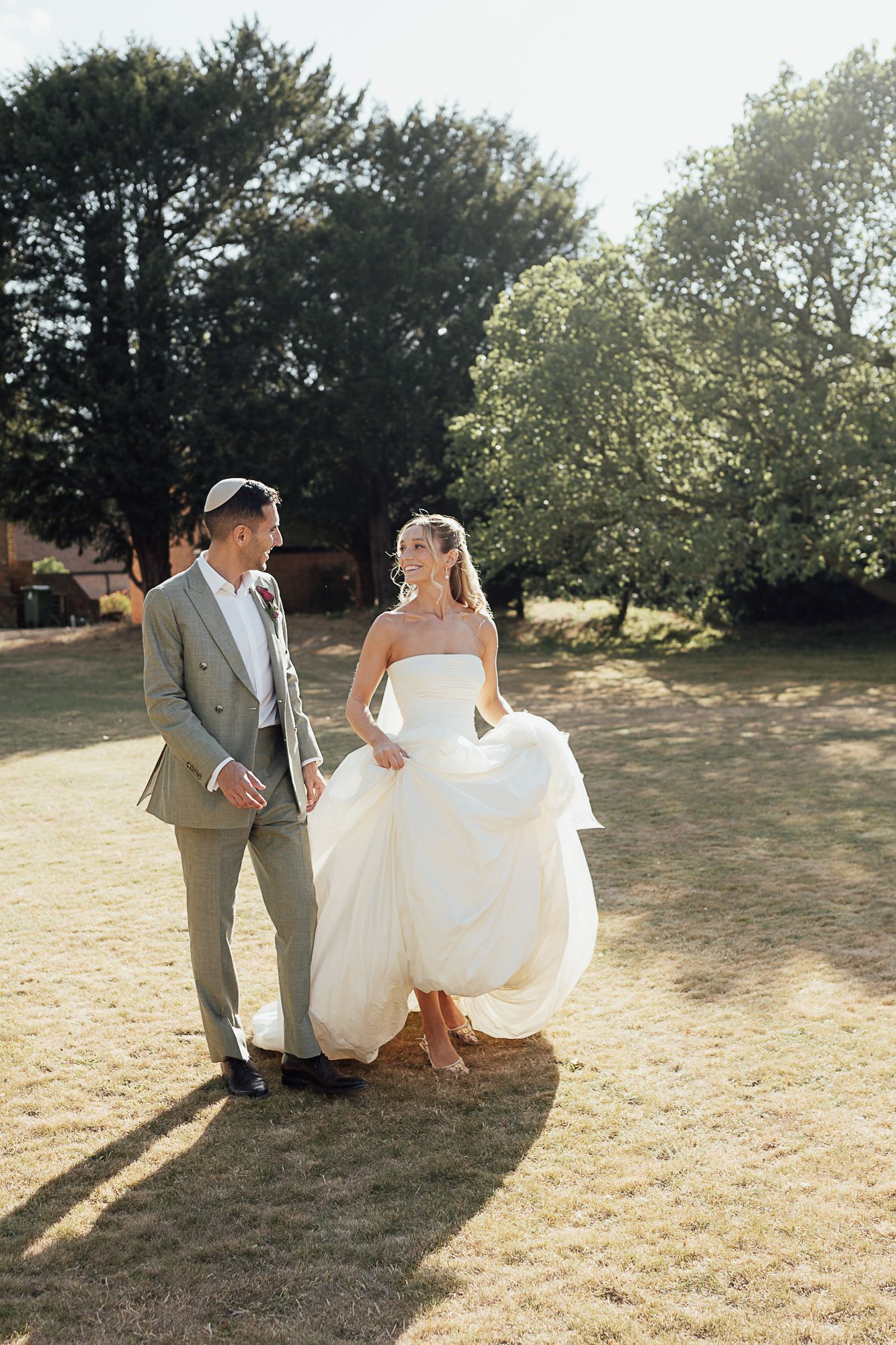 Bride in white strapless gown lifts dress while walking with groom in grey suit on sunny garden lawn.