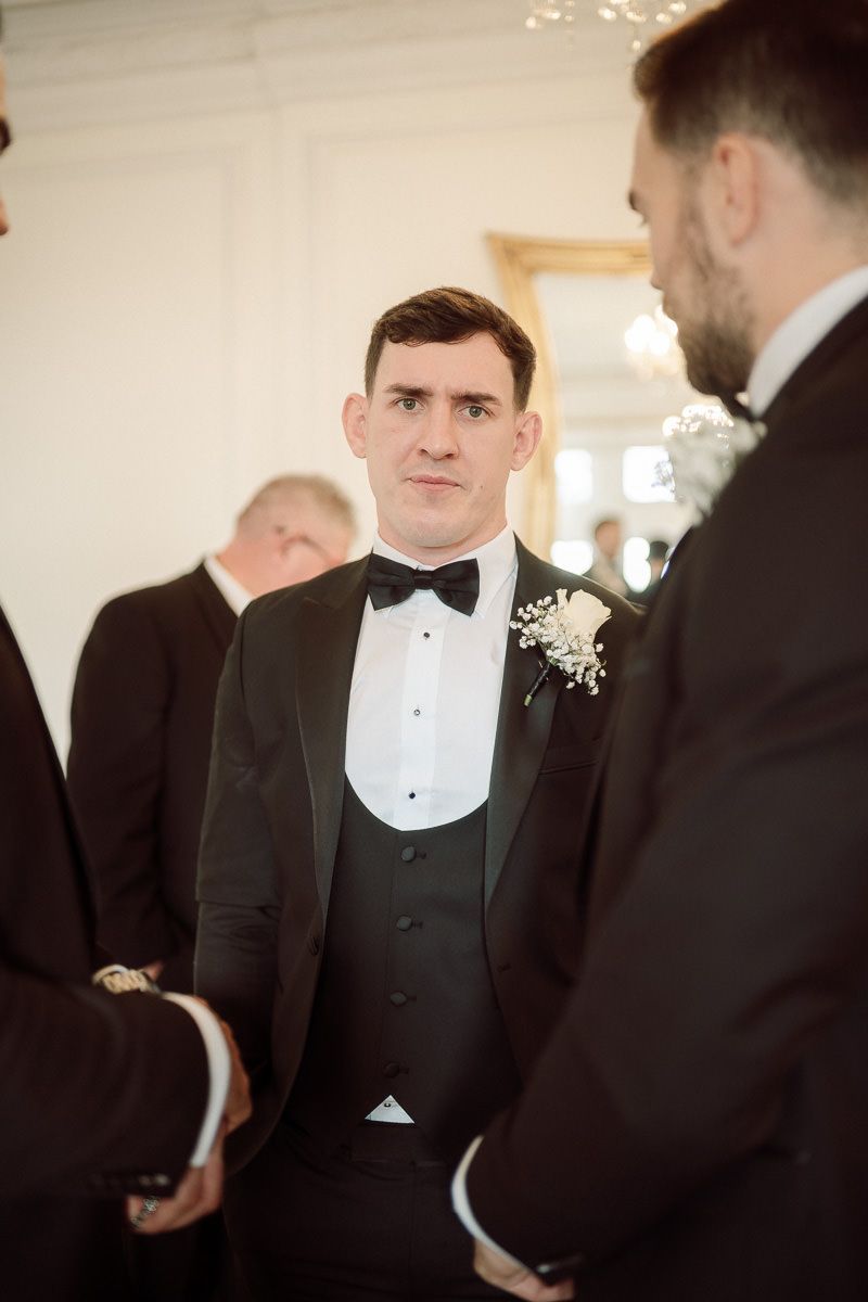 Groomsmen in black tuxedos and bow ties gather before a wedding ceremony.