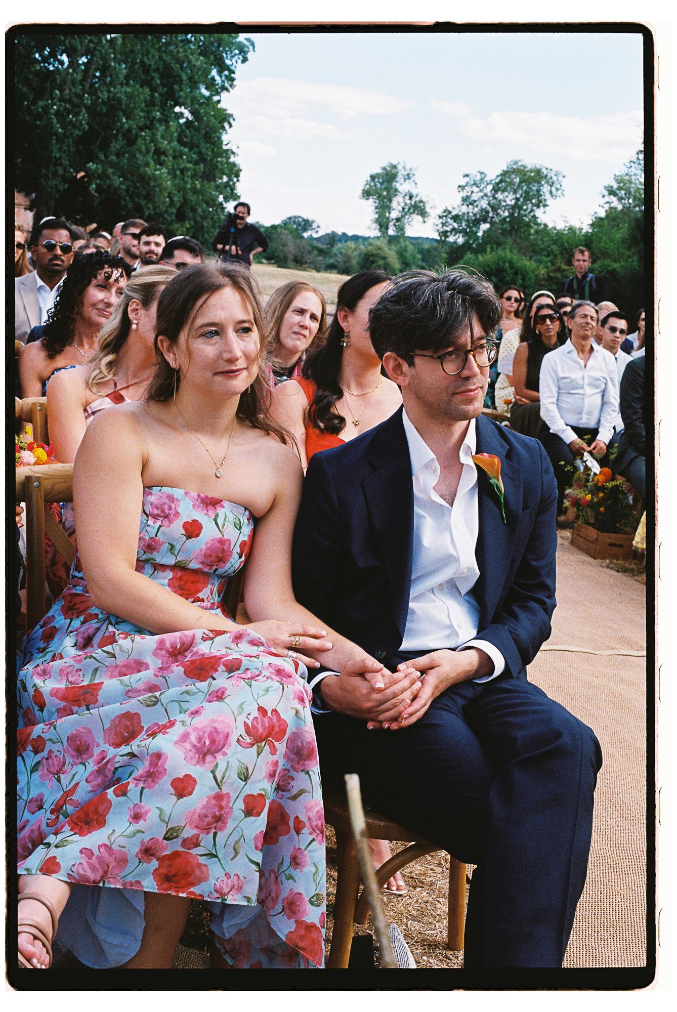 Couple in floral dress and navy suit seated at outdoor wedding ceremony with guests in background.