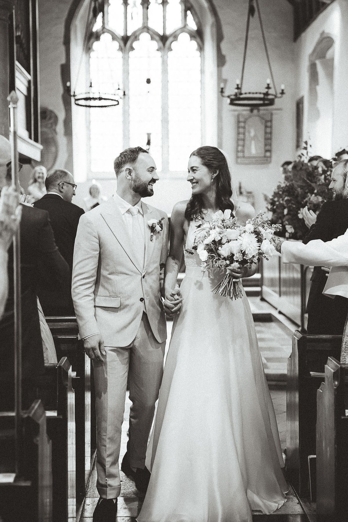 A romantic black and white wedding ceremony photo inside a beautiful church with stained glass windows and hanging chandeliers.