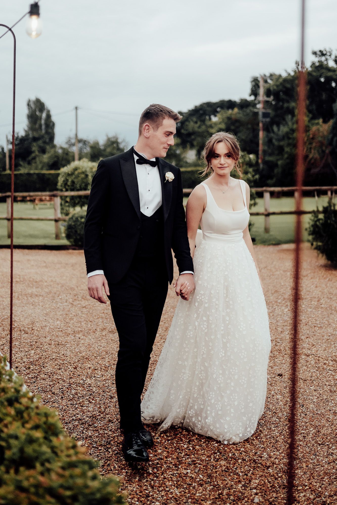A romantic wedding portrait of a couple walking together on a gravel path, the bride in white and groom in black tuxedo.