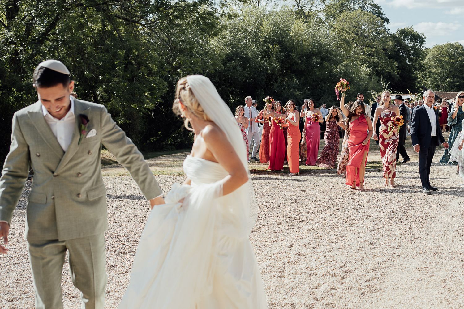 Bride and groom lead wedding party across gravel path outdoors, bridesmaids in pink and red dresses following behind.