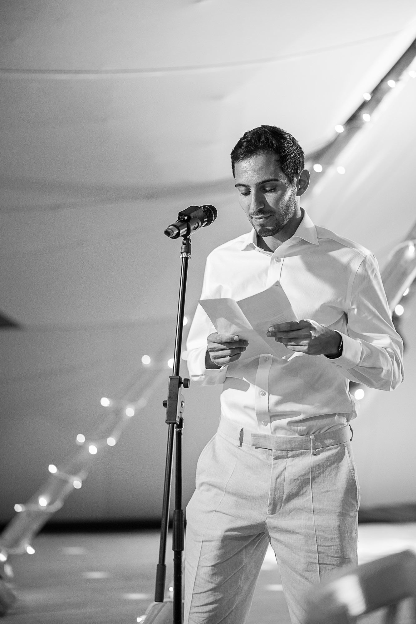 Man in white shirt reads notes at microphone stand during elegant wedding reception with twinkling lights.