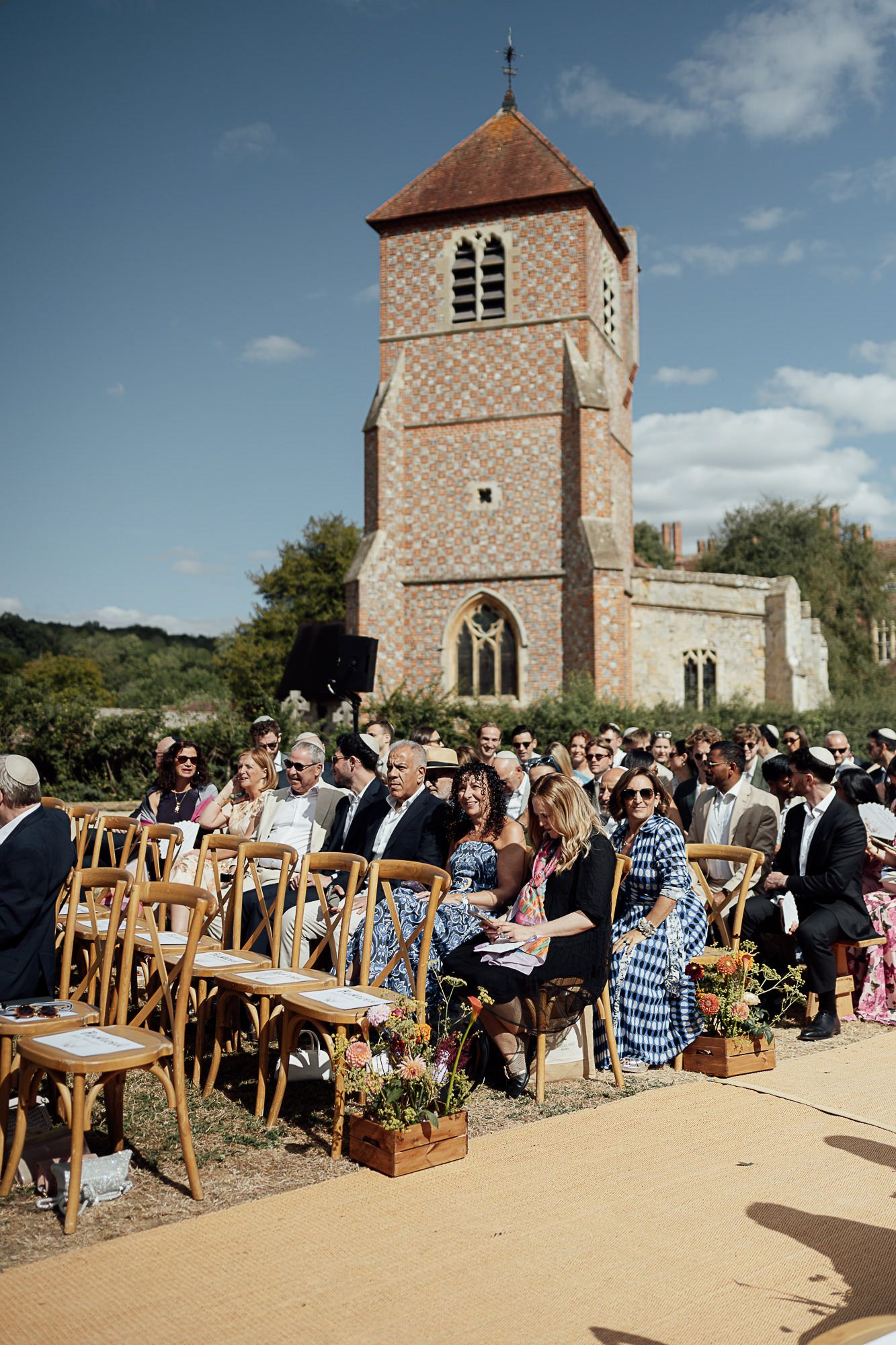 Guests seated in wooden chairs at an outdoor wedding ceremony beside a historic brick church tower on a sunny day.