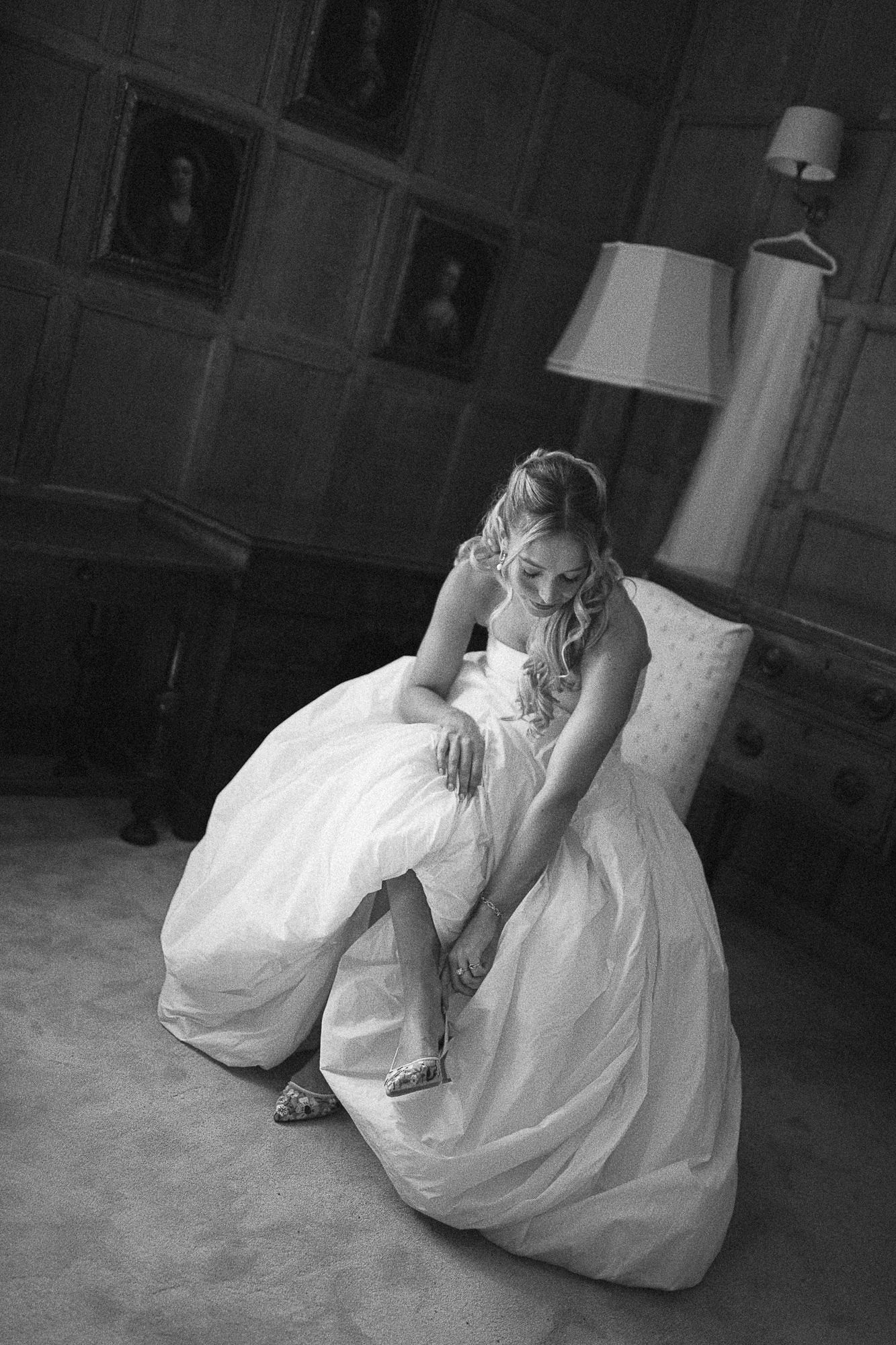 Black and white photo of a bride in a white gown sitting and fastening her wedding shoes in an elegant room.