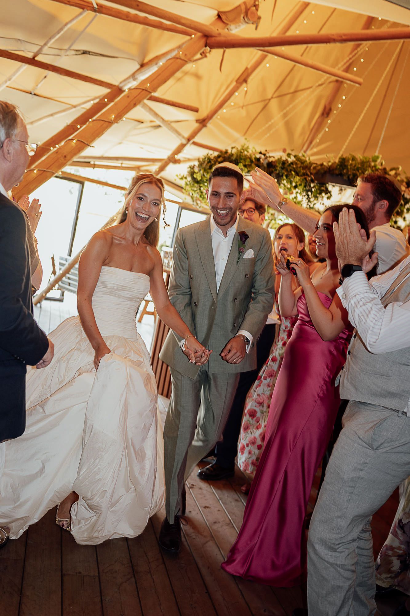 Bride and groom walk hand in hand under a tent as smiling wedding guests celebrate around them.