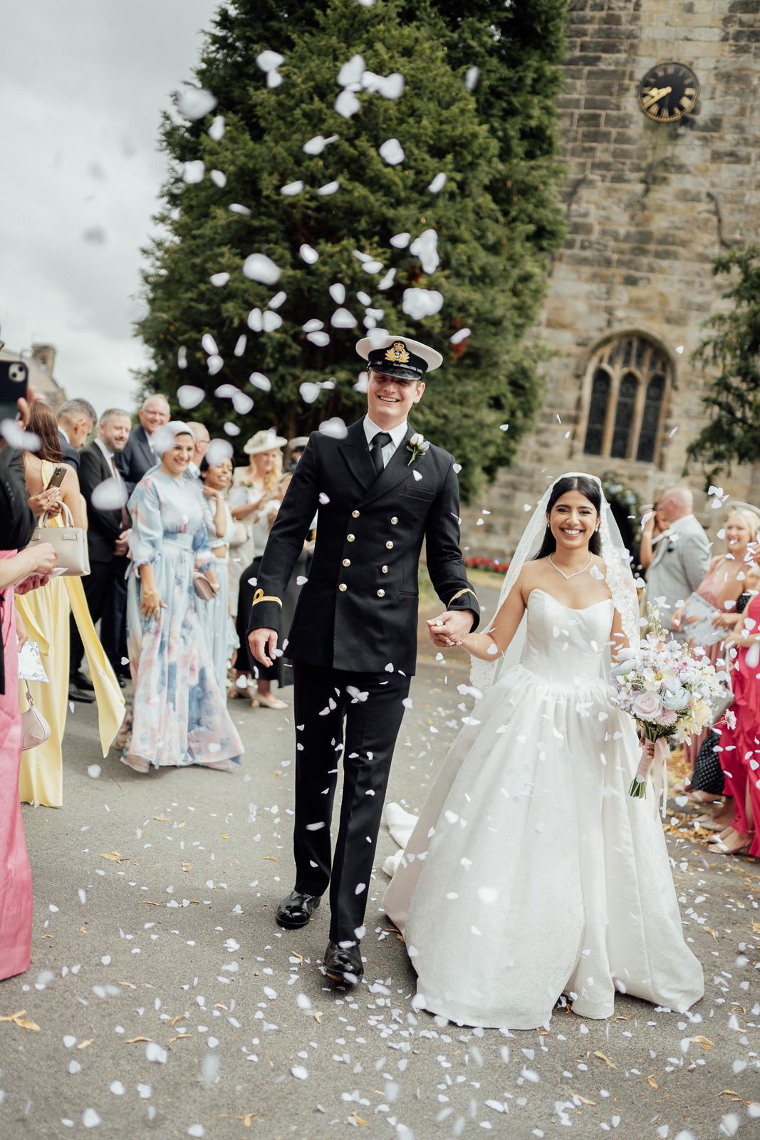 Newlywed couple walks through confetti shower outside historic stone church, groom in naval uniform, bride in white gown.