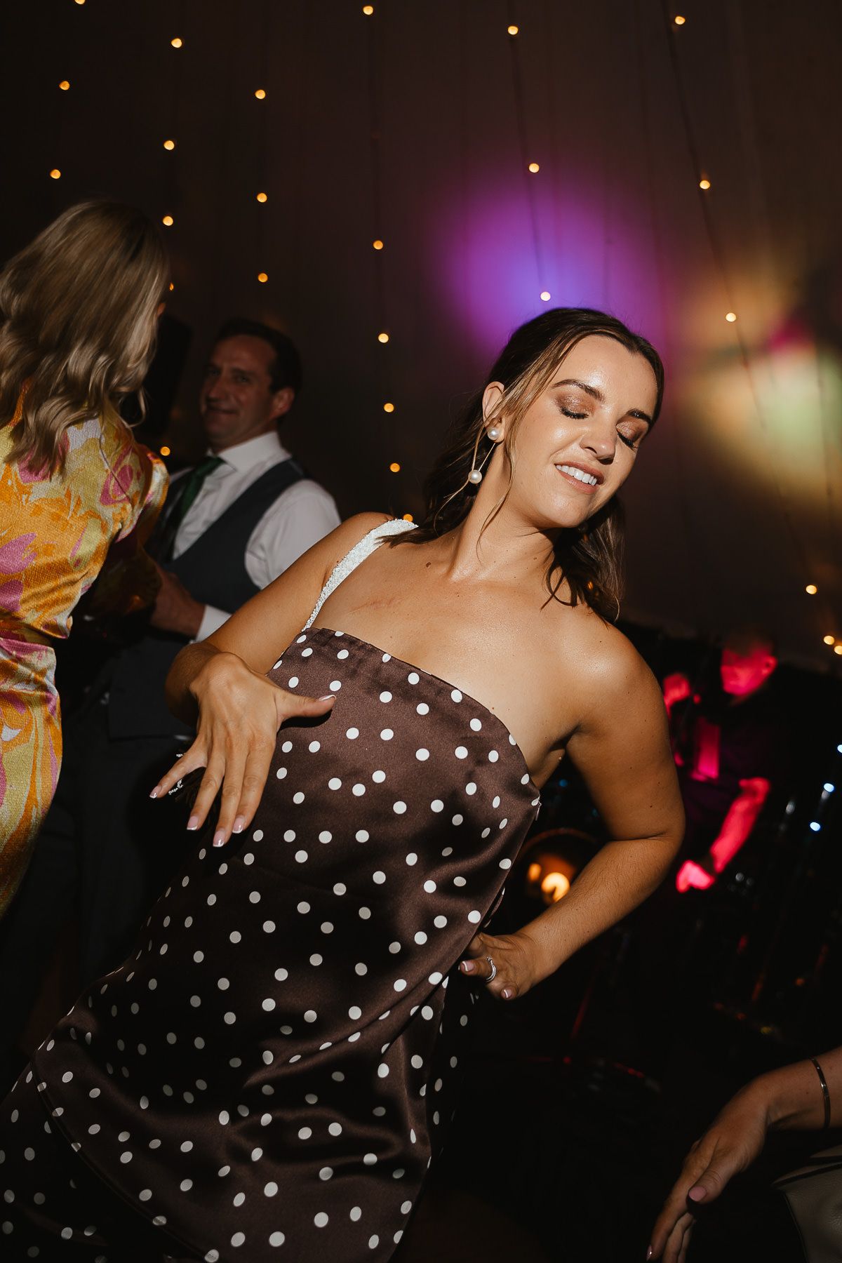A person in a brown polka dot dress dances expressively at a party with twinkling lights in the background.