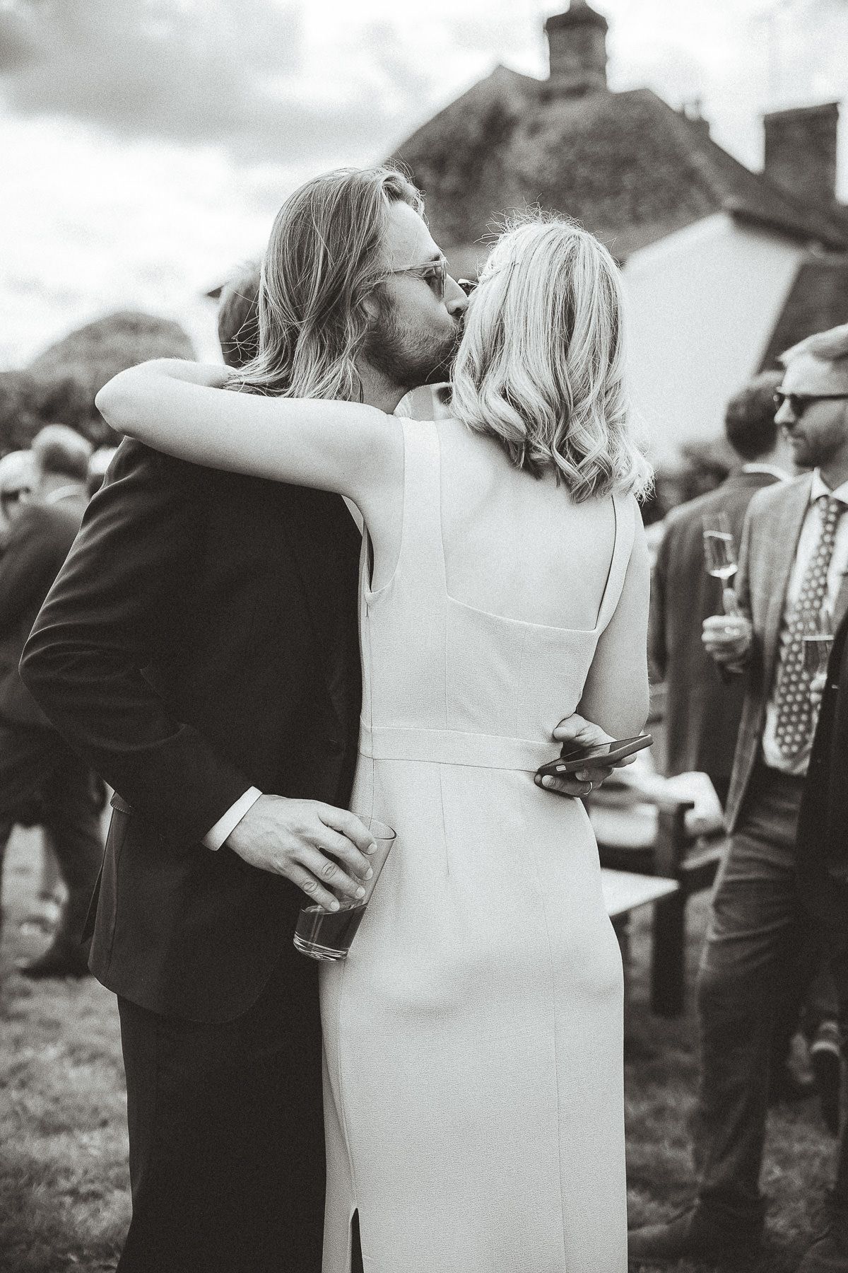 A couple in wedding attire share an intimate embrace during an outdoor reception with guests in the background.