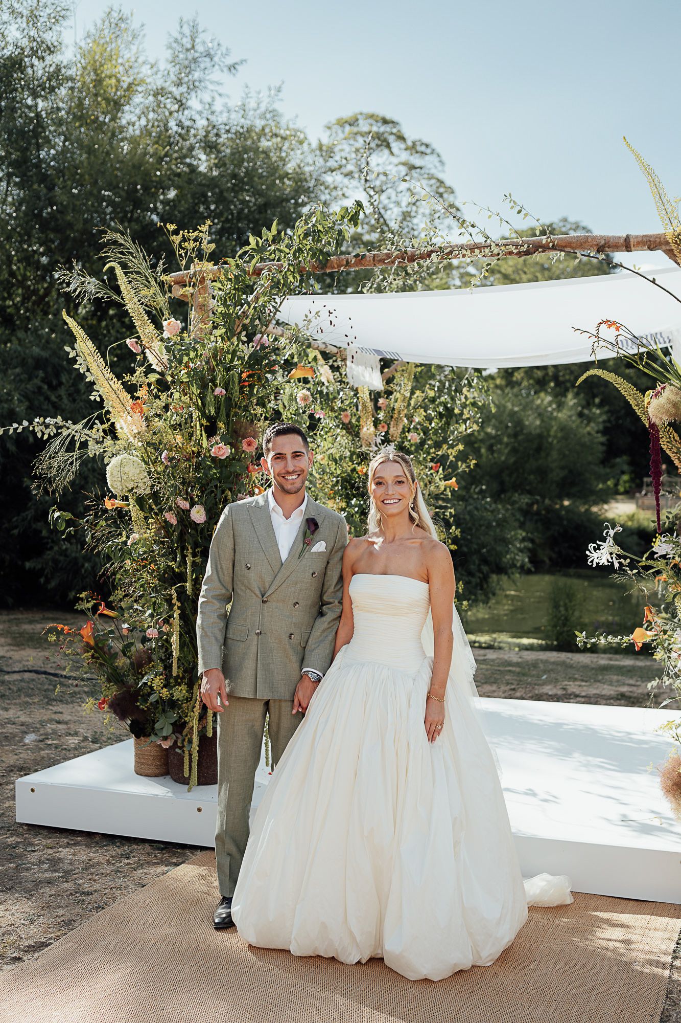 Bride in strapless white ballgown and groom in gray suit stand before floral arch at outdoor wedding ceremony.