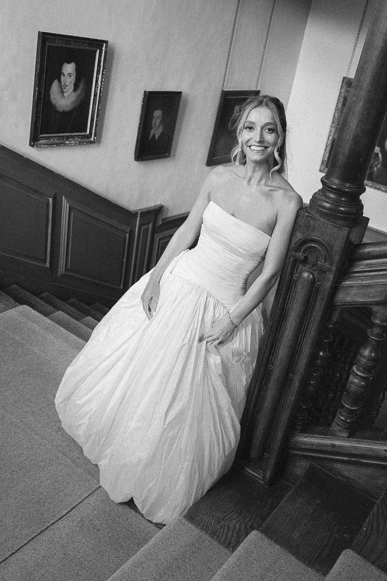 Smiling bride in strapless white ball gown poses on elegant wooden staircase with portrait paintings in black and white photo.