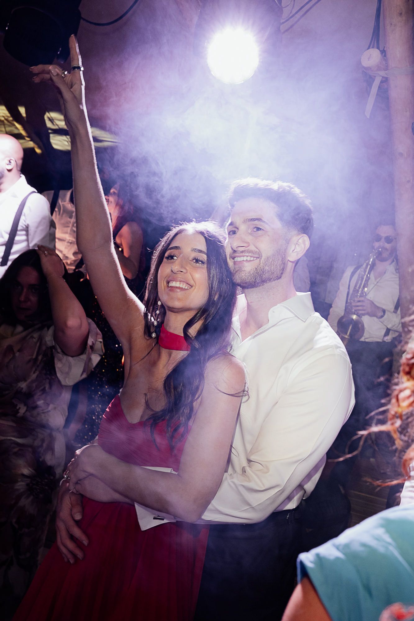 Couple dancing joyfully at a party, woman in red dress raises arm under bright stage lights with crowd behind them.