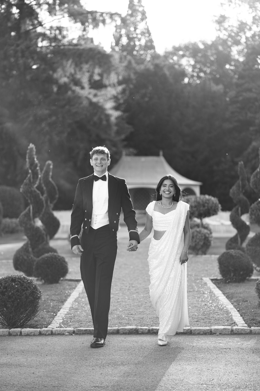 Bride in white saree and groom in tuxedo walking hand in hand through elegant garden in black and white photo.