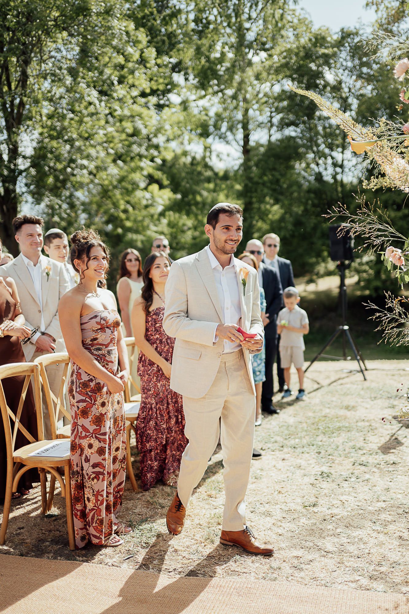 Groom in beige suit walks down outdoor wedding aisle lined with guests in summer attire amid lush greenery.