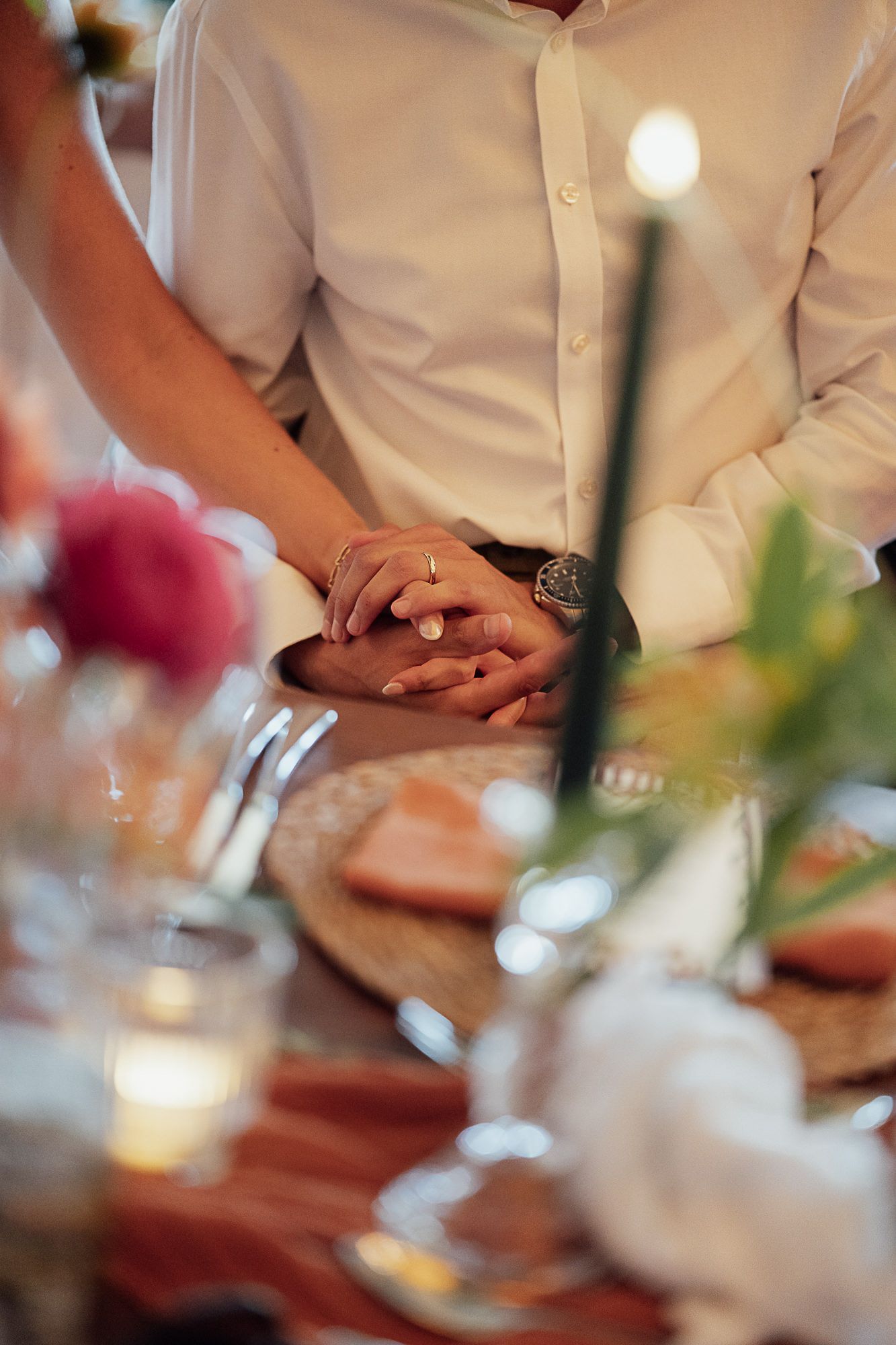 Couple holding hands at wedding reception table with candles, flowers, and elegant place settings in soft focus.