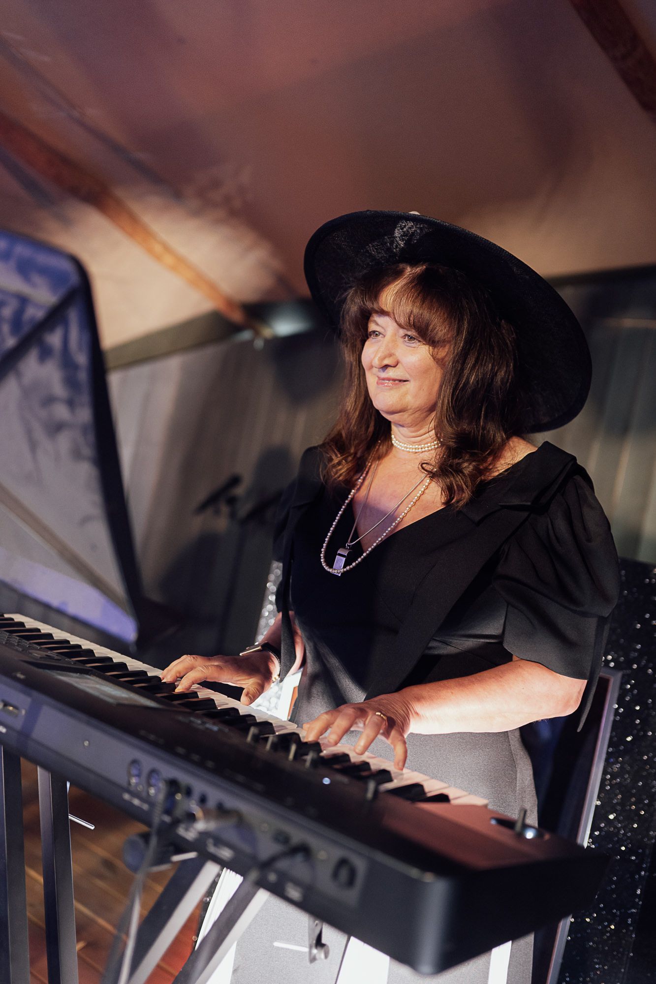 Woman in black hat and dress playing electronic keyboard piano at a live music performance event.