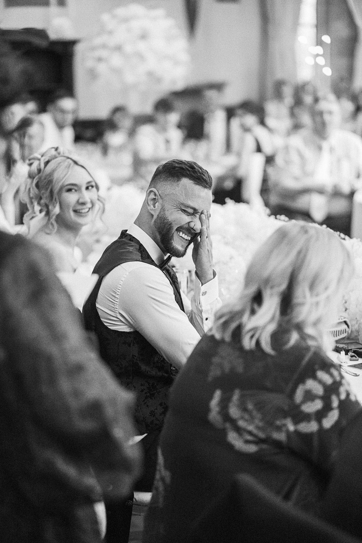 Black and white wedding reception photo of laughing groom in vest surrounded by smiling guests at elegant venue.