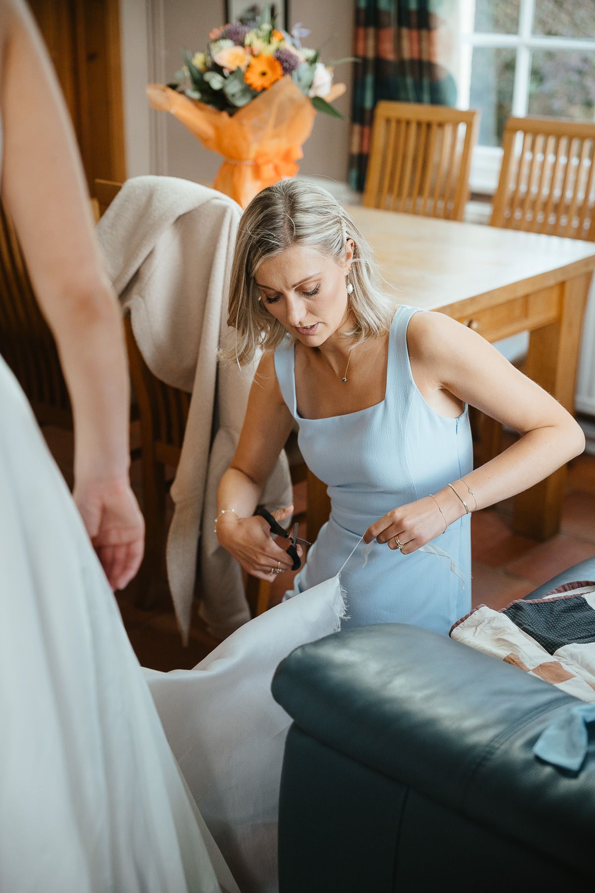 A person in a light blue dress helps adjust a white wedding gown in a dining room with a floral bouquet in the background.