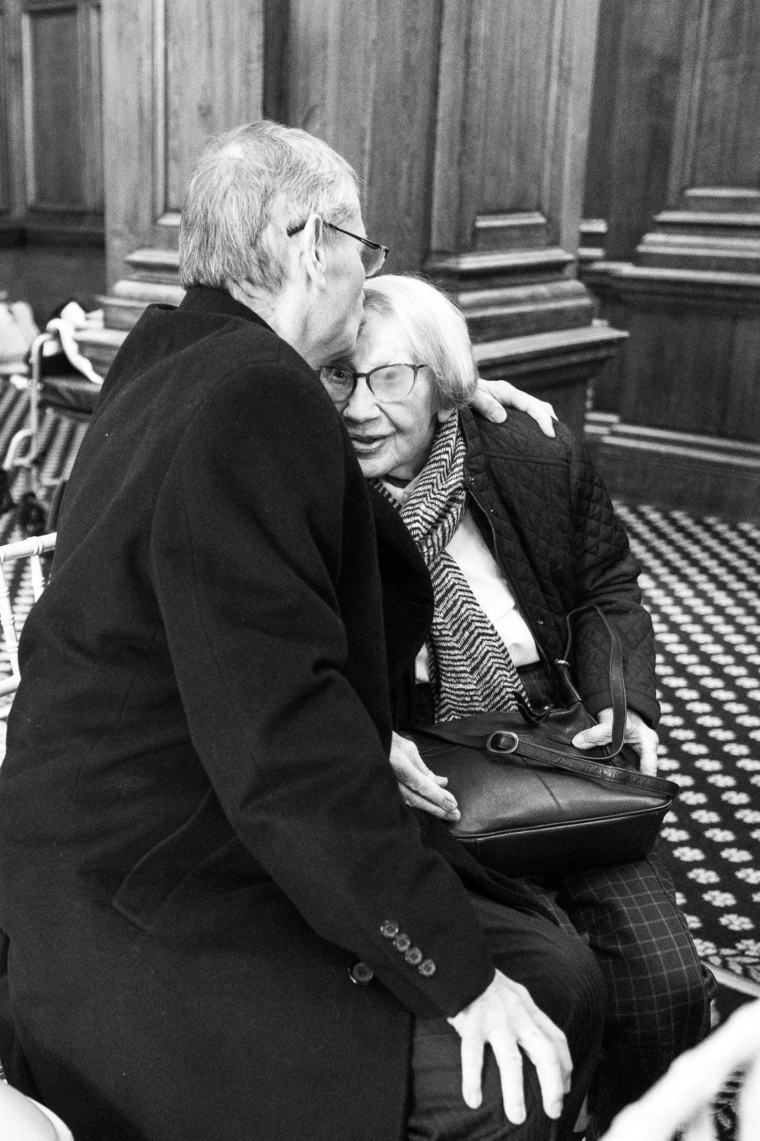 Intimate black and white shot of elderly couple sharing a tender moment in an elegant wood-paneled room.