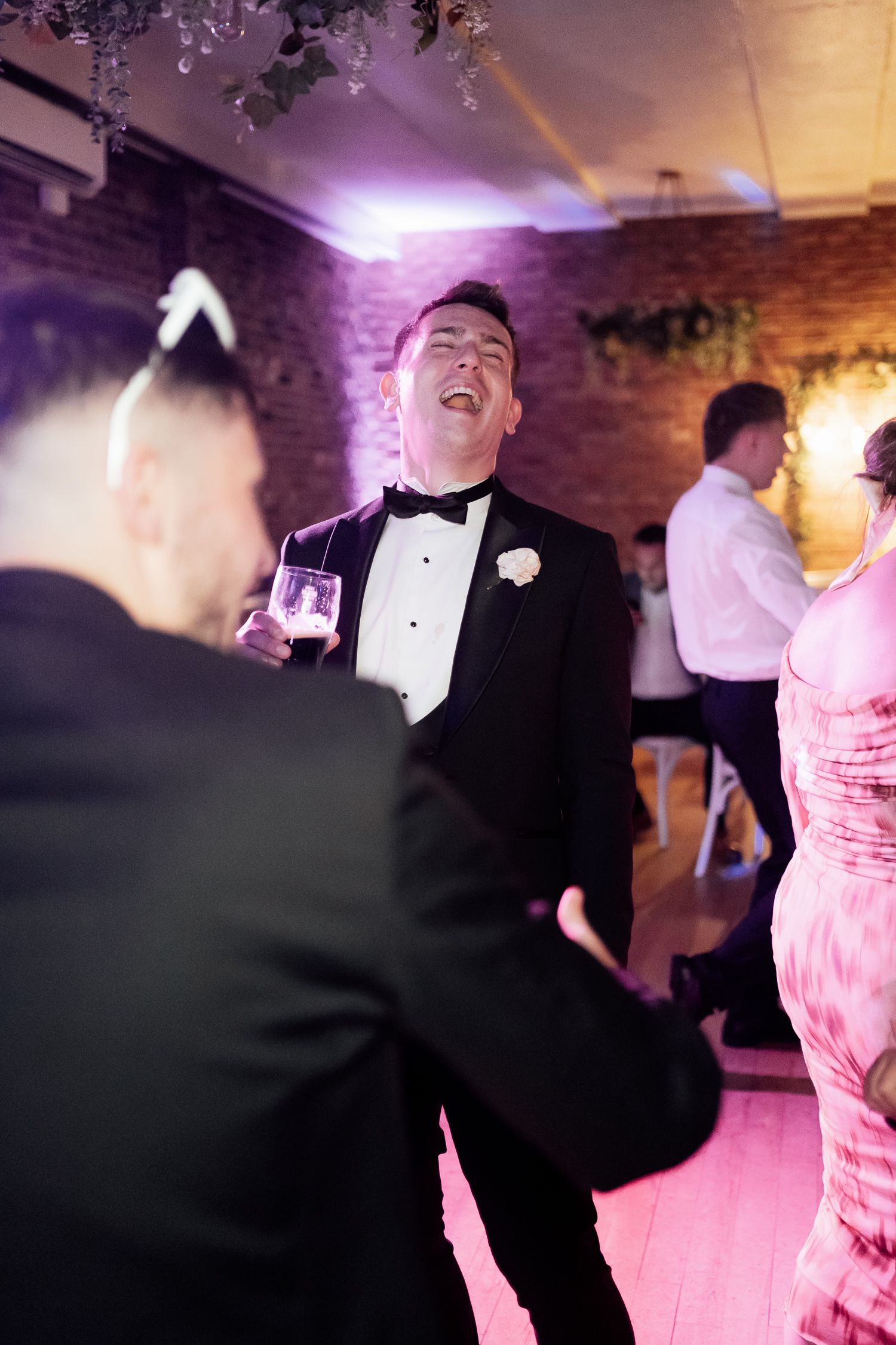 A person in a tuxedo laughing joyfully on a dance floor at a wedding reception with pink and purple lighting.