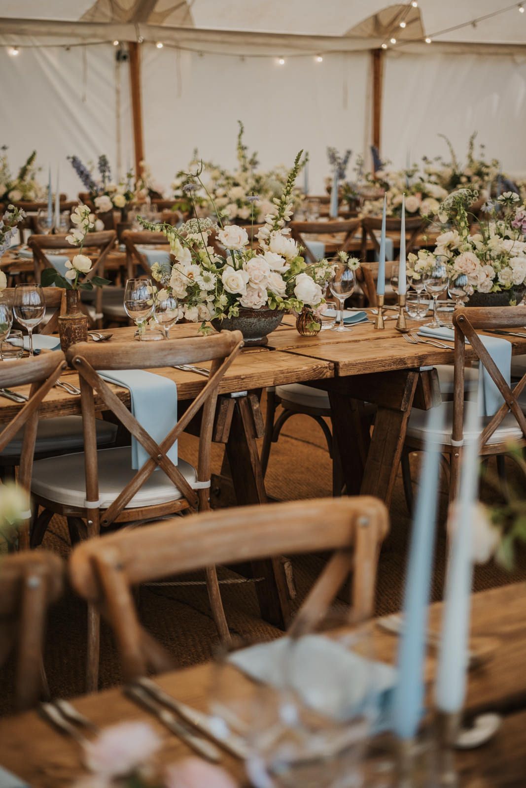 Elegant wedding reception table setup with rustic wooden farm tables, white floral centerpieces and crossback chairs under tent.