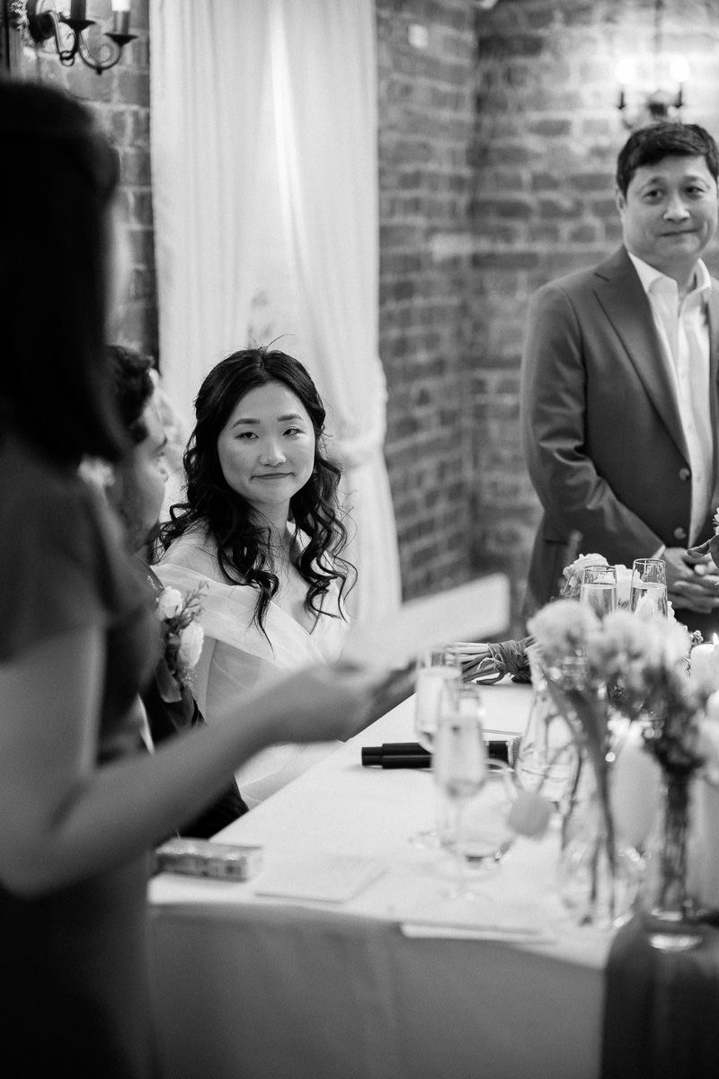 Wedding guests seated at elegant tables with white linens and floral centerpieces in a brick-walled venue.