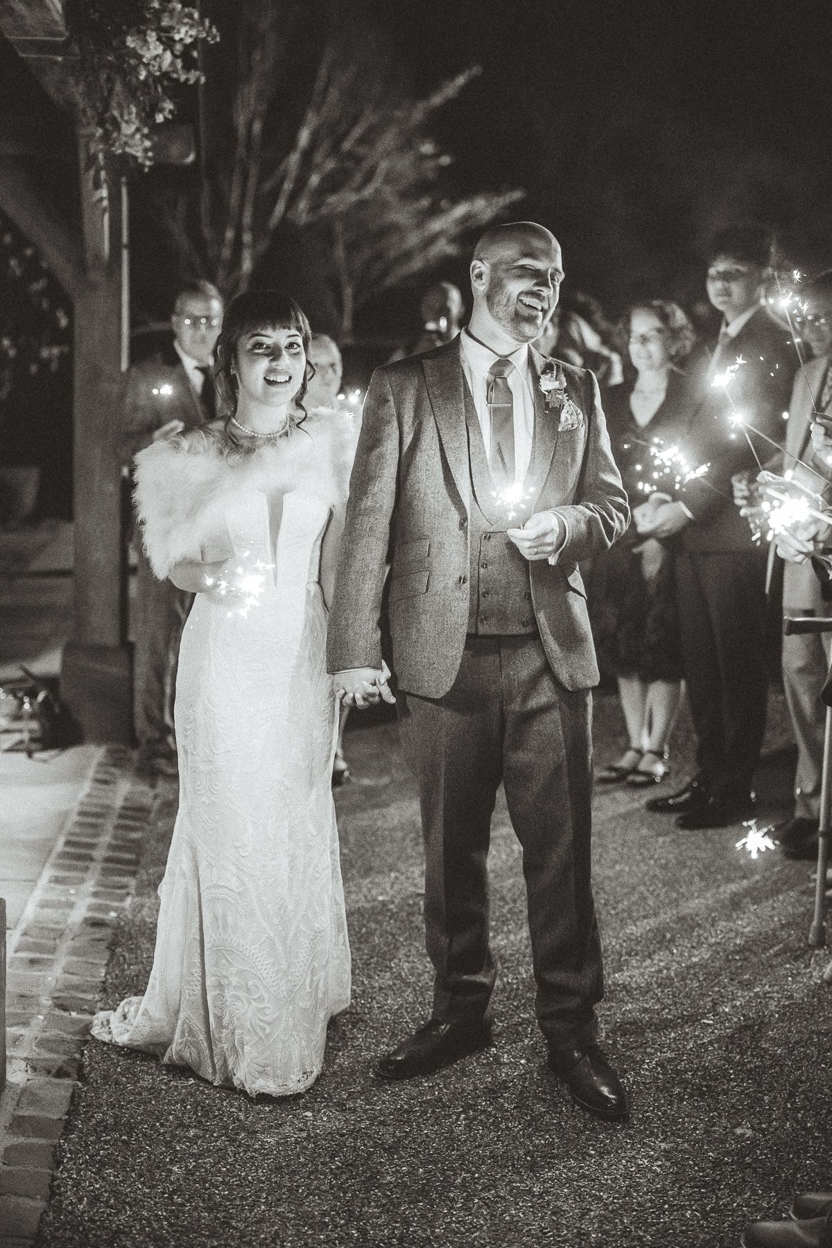 A black and white nighttime wedding celebration with sparklers illuminating a joyful couple in formal attire.