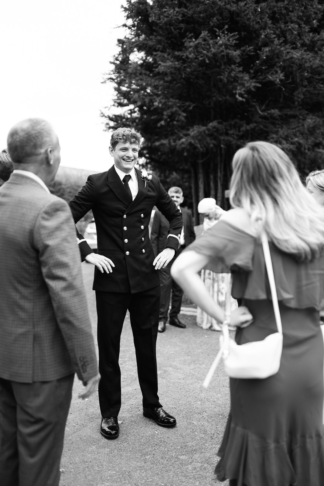 Black and white photo of a smiling man in a dark suit chatting with guests outdoors at a formal event.