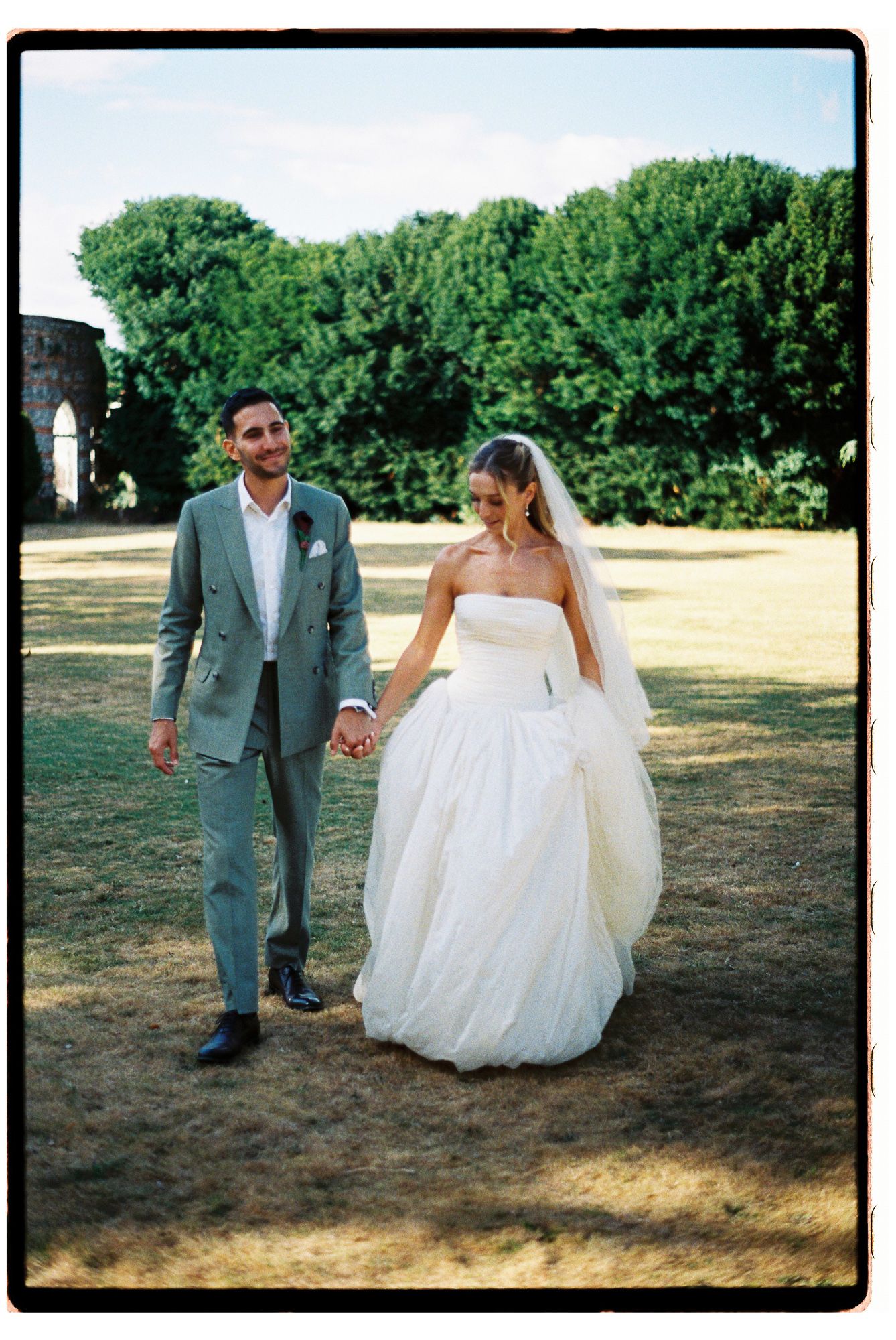 Bride and groom holding hands walking outdoors, she in white strapless gown with veil, he in sage green suit.