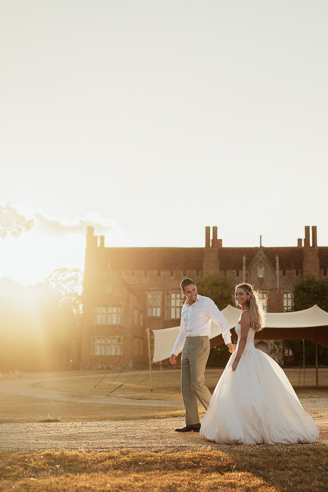Bride and groom walking hand in hand at golden sunset outside a historic brick venue.