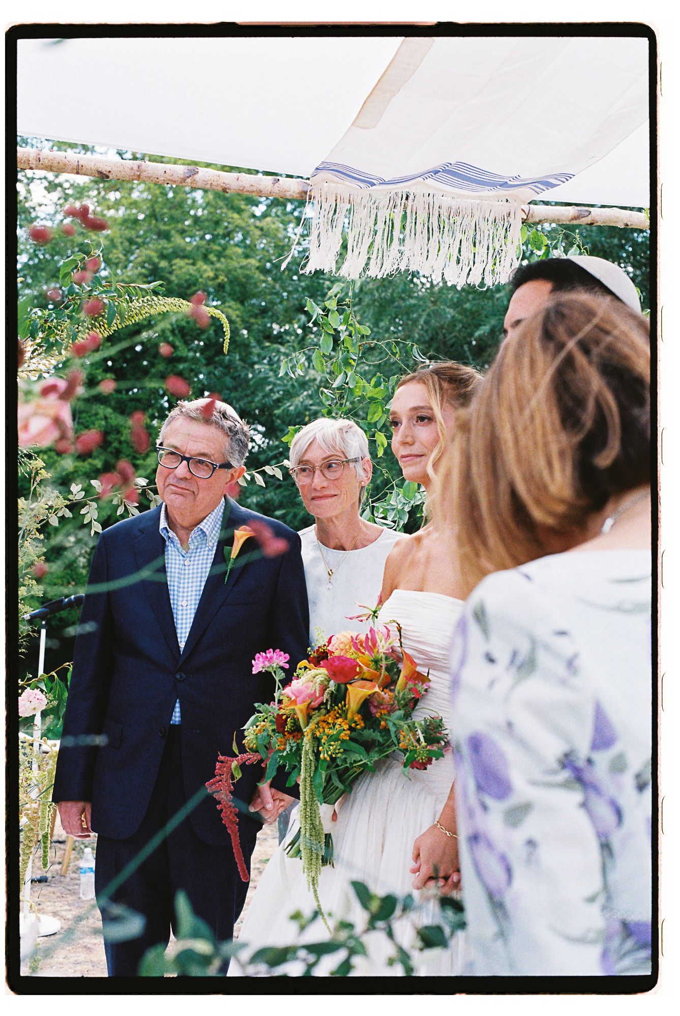 Bride holding colorful bouquet stands under chuppah with family during outdoor Jewish wedding ceremony.